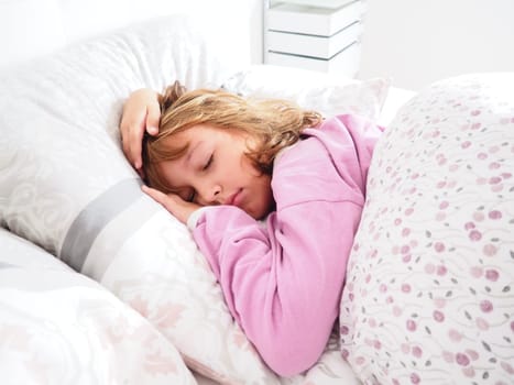 A beautiful caucasian girl of 8 years old with blond hair, dressed in pink pajamas, sleeps on a bed with a fluffy blanket, hugging a pillow. Soft light of the morning sun streams through the window