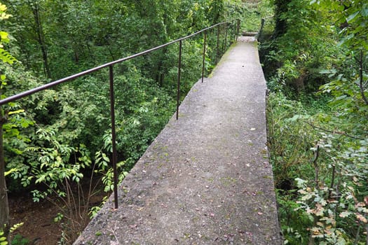 Banja Koviljacha, Serbia, Loznica, Mount Guchevo. An old narrow concrete bridge across a mountain river with a metal fence. Thickets of bushes, ivy, vines, trees around the bridge. Walk in nature