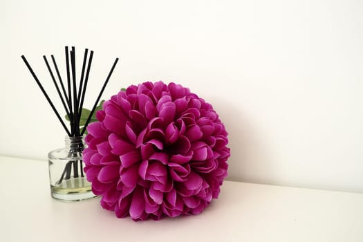 Arrangement with incense sticks, essential oil in a glass vase and a bright pink-lilac terry artificial flower in the interior of a white room. White background.