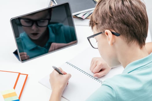 Top view of young preteen boy pupil wearing T-shirt, glasses, sitting at white desk near notepad in front of tablet, doing homework, writing on notebook with left hand. Distant learning, technology.