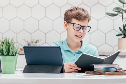 Portrait of young preteen smiling boy wearing blue T-shirt, glasses, sitting at desk near notepads, looking at open black book near tablet, having online lesson, reading text. E-learning, technology.