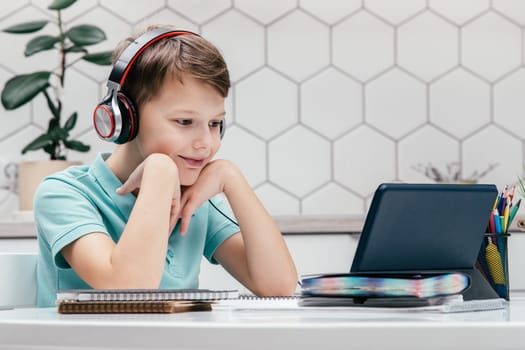 Portrait of young preteen interested boy wearing headphones, sitting at desk near notebooks, looking at screen of tablet, having online lesson, listening, leaning on hands. E-learning, technology.