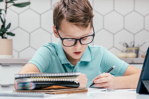 Portrait of preteen focused boy pupil wearing T-shirt, glasses, sitting at desk near notebooks, tablet, doing homework, writing on notebook, holding pen with left hand. Distant learning, education.