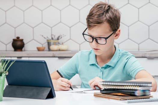 Portrait of preteen serious boy pupil wearing blue T-shirt, glasses, sitting at desk, looking at screen of tablet, doing homework, writing on notebook with left hand. Distant learning, education.