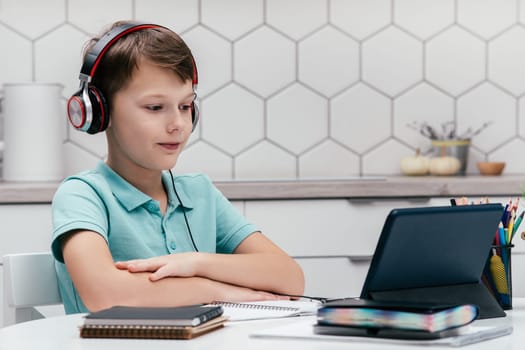 Portrait of young preteen focused boy wearing T-shirt, headphones, sitting at desk near notebooks, looking at screen of tablet, having online lesson, listening. Remote education, distance learning.