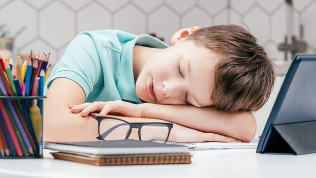 Portrait of young preteen tired boy wearing blue T-shirt, lying sleeping on hands on desk near notebooks, glasses, color pencils, tablet at home. Distant learning, remote education, technology.