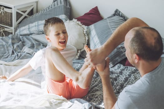 Father plays with his son 7-10 on bed. Dad tickles kids feet. Family having fun