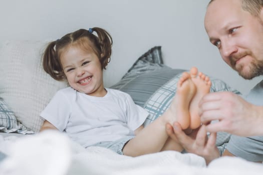 Father plays with little daughter 2-4 on bed. Dad tickles kids feet. Family having fun