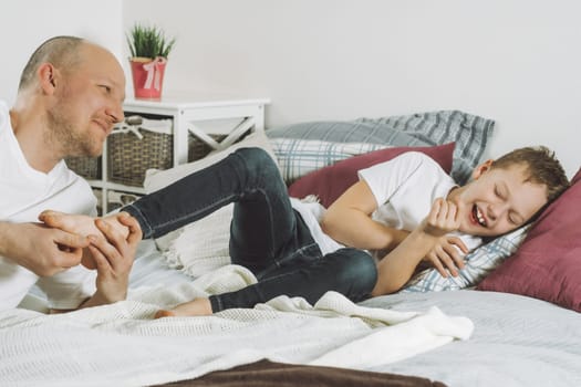 Father plays with his son 7-10 on bed. Dad tickles kids feet. Family having fun