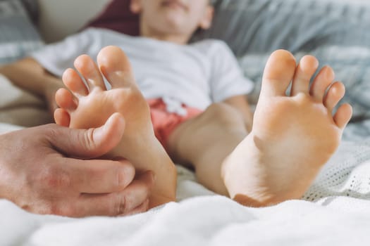 Dad tickles feet of kid. Boy lies on bed and plays with his father. Family having fun at home