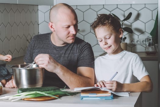 Father whips omelet and helps his son do his homework in the kitchen. Man doing household chores.