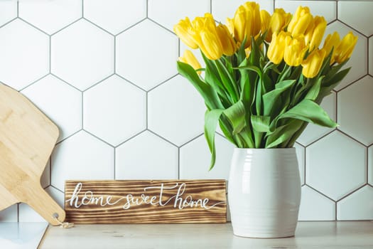 Interior shot. Home sweet home handwritten sign on kitchen countertop next to yellow tulip bouquet in vase and cutting board on hexagonal white tile background