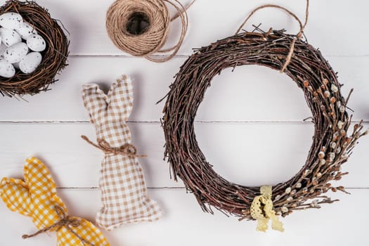 Easter crafting woven willow wreath with tape bow. White planking wall background. Eggs nest and toy rabbits invitation. Tradition festive seasonal decoration step by step. Workshop ornament. Top view