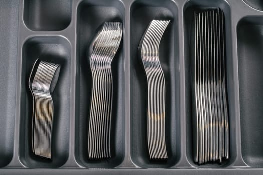Kitchen cabinet open drawer with black cutlery organizer tray with silver forks, spoons and knives. Kitchen storage organization system. Top view