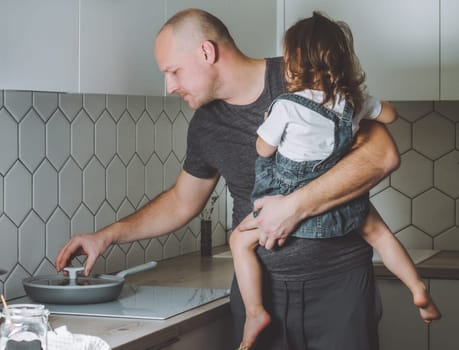 Father is preparing dinner in kitchen with his daughter in his arms. Man doing household chores.