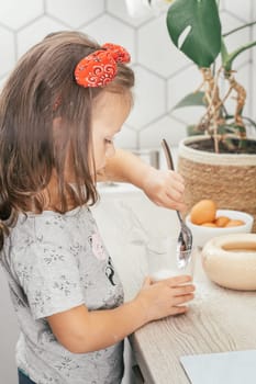 Little dark-haired girl 3 years old in red headband bakes apple pie in kitchen. Child puts sugar in measuring cup. Children help on household chores. Kid cooking food. Vertical