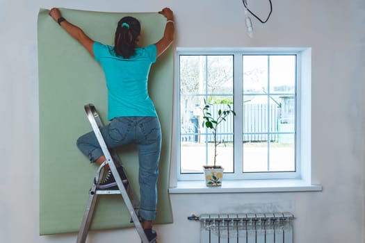 Woman in jeans and turquoise T-shirt standing on stepladder and holding piece of wallpaper. Home renovation and improvement.