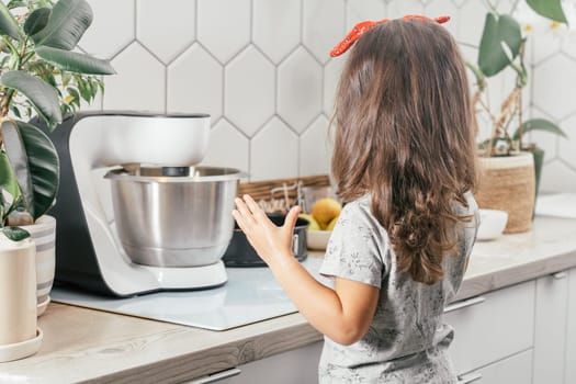 Little dark-haired girl 3 years old in red headband bakes apple pie in kitchen. Child stands near planetary mixer. Children help on household chores. Kid cooking food. Back view