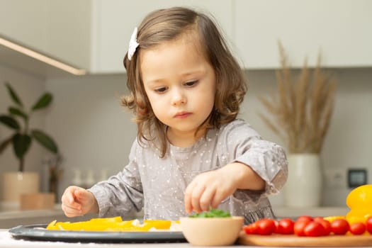 Cute little girl 2-4 in gray dress cooking pizza in kitchen, kid arranges ingredients on pizza base