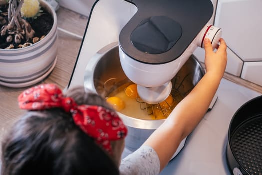 Little dark-haired girl 3 years old in red headband bakes apple pie in kitchen. Child turns on planetary mixer. Children help on household chores. Kid cooking food. Top view