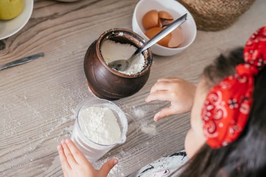 Hands of little girl in red headband bake apple pie in kitchen. Child puts flour in measuring cup. Children help on household chores. Kid cooking food. Top view