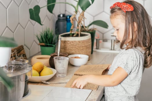 Dark-haired girl 3 years old in red headband bakes apple pie in kitchen. Child stands near table with ingredients, cooking utensils and recipe book. Children help on household chores. Kid cooking food