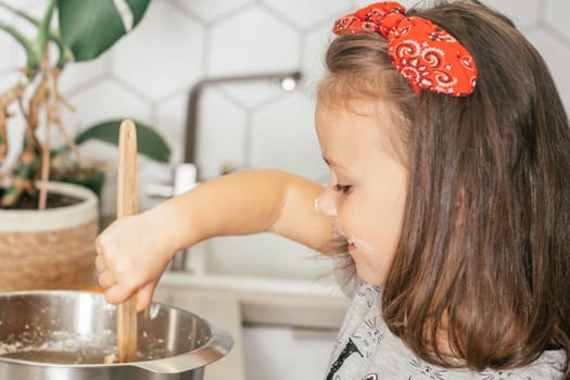 Little dark-haired girl 3 years old in red headband bakes apple pie in kitchen. Child mixes dough in bowl. Children help on household chores. Kid cooking food
