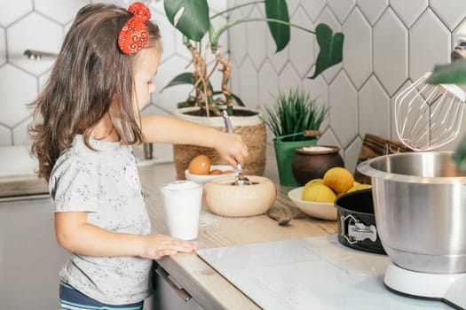 Little dark-haired girl 3 years old in red headband bakes apple pie in kitchen. Child puts sugar in measuring cup. Children help on household chores. Kid cooking food