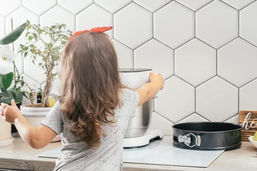 Little dark-haired girl 3 years old in red headband bakes apple pie in kitchen. Child turns on planetary mixer. Children help on household chores. Kid cooking food. Back view