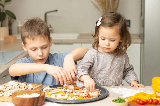 Cute little girl 2-4 in gray dress and boy 7-10 in T-shirt cooking pizza together in kitchen, brother and sister cooking