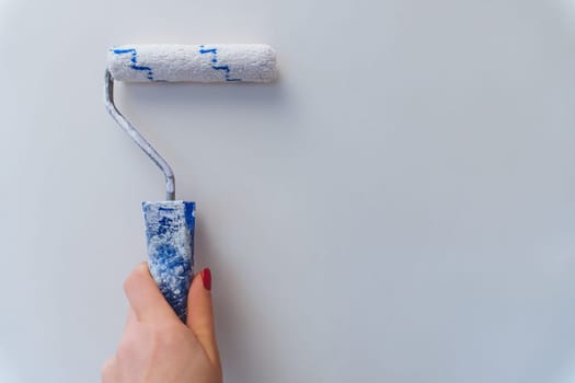 Female hand with red manicure holds roller and paints wall white. Home renovation and improvement.