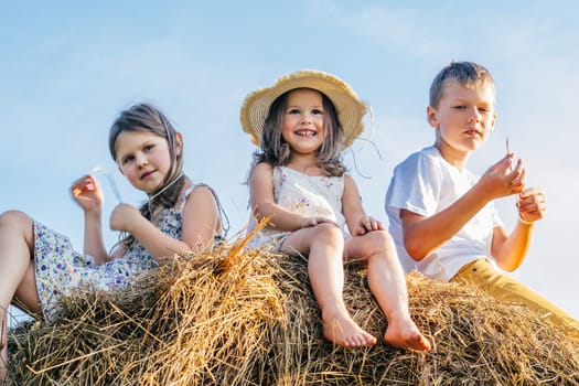 Portrait of three kids boy and two girls sitting on haystack in summer field. Light sunny day. Cheerful and enjoy concept. Low angle. Blue sky on background. Girl wearing straw hat and smiling.