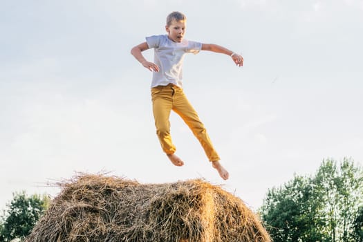Portrait of barefoot boy in T-shirt and trousers jumping on haystack in field. Light sunny day. Cheerful and enjoy concept. Fresh air in countryside. Trees on background. Outdoor activity