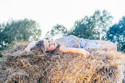 Portrait of little blond girl in dress alone lying and relaxing on haystack in field. Light sunny day. Rest and calm concept. Fresh air in countryside. Side view. Trees and clear sky on background.