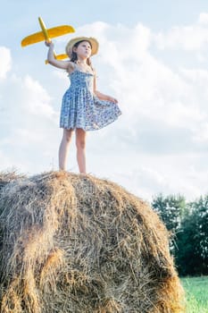 Portrait of little girl playing, standing on haystack in field. Flying throwing yellow toy airplane. Light sunny day. Purposefully concept. New beginning, looking to future, travelling. Low angle