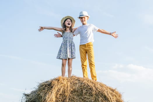 Portrait of children boy and girl staying and hugging, singing songs, waving hands playing the ape on haystack in field. Light sunny day. Kids wearing straw hats. Young artists outdoors. Low angle