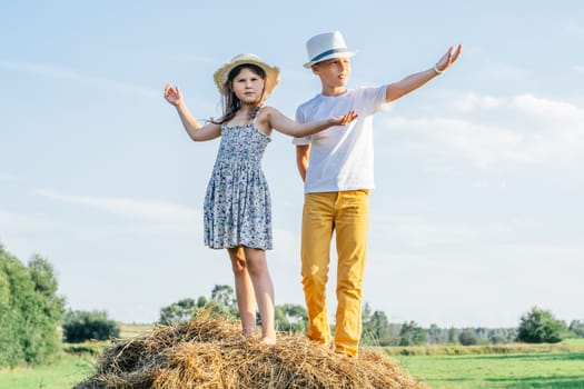 Portrait of children boy and girl staying spreading and waving arms, playing the ape on haystack in field. Light sunny day. Trees and clear bright sky on background. Kids wearing straw hats and posing