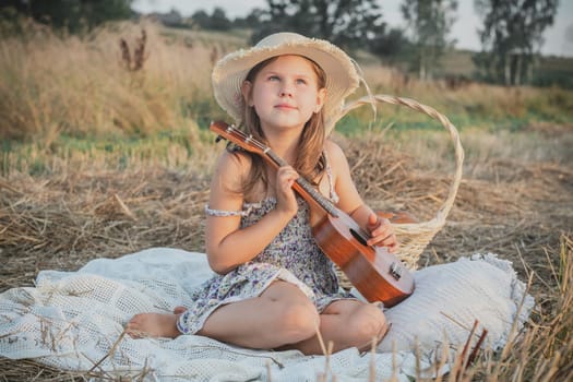 Portrait of smiling girl sitting on blanket in dry hay field, having picnic, learning playing guitar ukulele. Resting and relaxing time. Basket near kid. Music concept