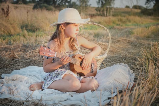 Portrait of girl sitting on blanket in dry hay field, having picnic, learning playing guitar ukulele. Resting and relaxing time. Basket near kid. Side view. Music concept