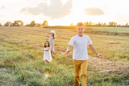 Portrait of three children playing game of catch, jumping and running on dry grass hay field paths in sunset. Forest and meadow on background. Looking around. Cloudy sunny sky. Haying time