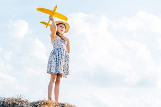 Portrait of little girl in hat and dress playing flying yellow toy airplane, standing on haystack. Taking aim in sky. Light sunny day. Having adventure. Clear sky with clouds on background. Low angle