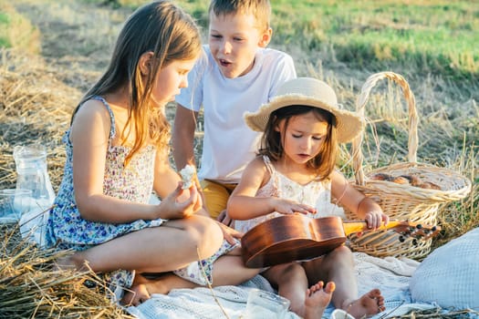 Portrait of joyful boy and two girls sitting on blanket in field, having picnic, eating bread. Relaxing time. Playing guitar, learning playing ukulele, talking, laughing. Light sunny day