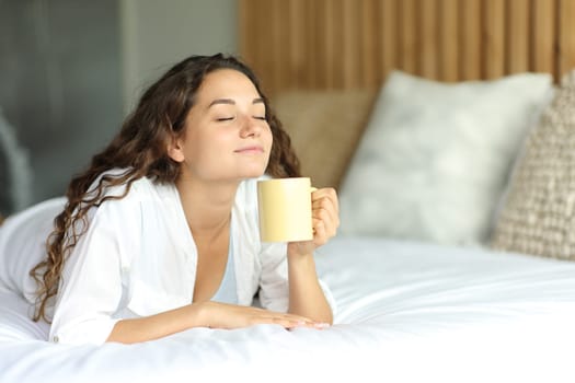 Happy woman lying on a bed enjoying a cup of coffee