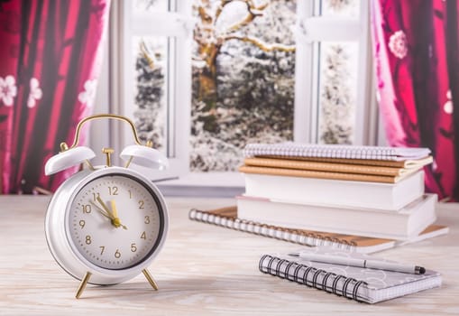 Winter semester, time management concept. Alarm clock with book, notenooks with open window in background