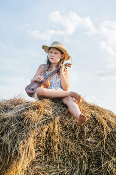 Nice little girl sit at top of haystack dangling legs and play ukulele. Carefree child resting on hayrick, from below view. Outdoor walking. Beautiful sky. Summer vacation, countryside lifestyle.