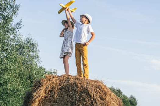 Little girl and boy together launch yellow toy airplane standing at top of haystack. Children on hayrick from below view. Freedom. Beautiful blue sky. Summer vacation, countryside lifestyle.