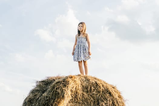 Pretty little girl stand at top of haystack looking away. Full length photo, from below view. Resting on hayrick. Outdoor walking. Beautiful blue sky. Freedom. Summer vacation, countryside lifestyle.