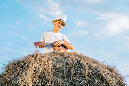 Handsome school aged boy sitting at top of haystack play ukulele. Carefree child resting on hayrick, from below view. Outdoor walking. Beautiful blue sky. Summer vacation, countryside lifestyle.