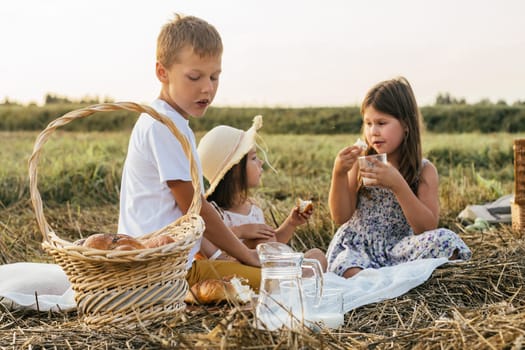 Happy friends talk, eat buns and drink milk sitting on blanket on field. Portrait of little children resting on grass, side view. Food basket for picnic. Summer vacation, countryside lifestyle.