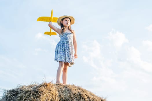 Serious little girl in hat stand at top of haystack and launch yellow toy airplane. Child on hayrick from below view. Outdoor walking. Beautiful blue sky. Summer vacation, countryside lifestyle.